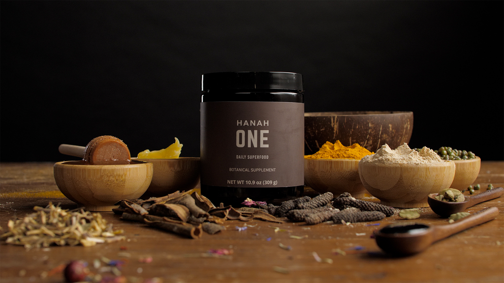 Jar of HANAH ONE, a botanical supplement, surrounded by its natural ingredients in wooden bowls and spoons.