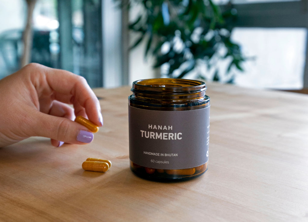Open jar of HANAH Turmeric on a wooden table with a hand holding a capsule next to it