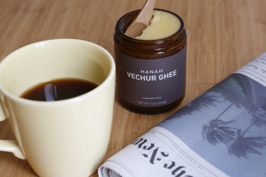 Open jar of HANAH Vechur Ghee next to a mug of coffee and newspaper