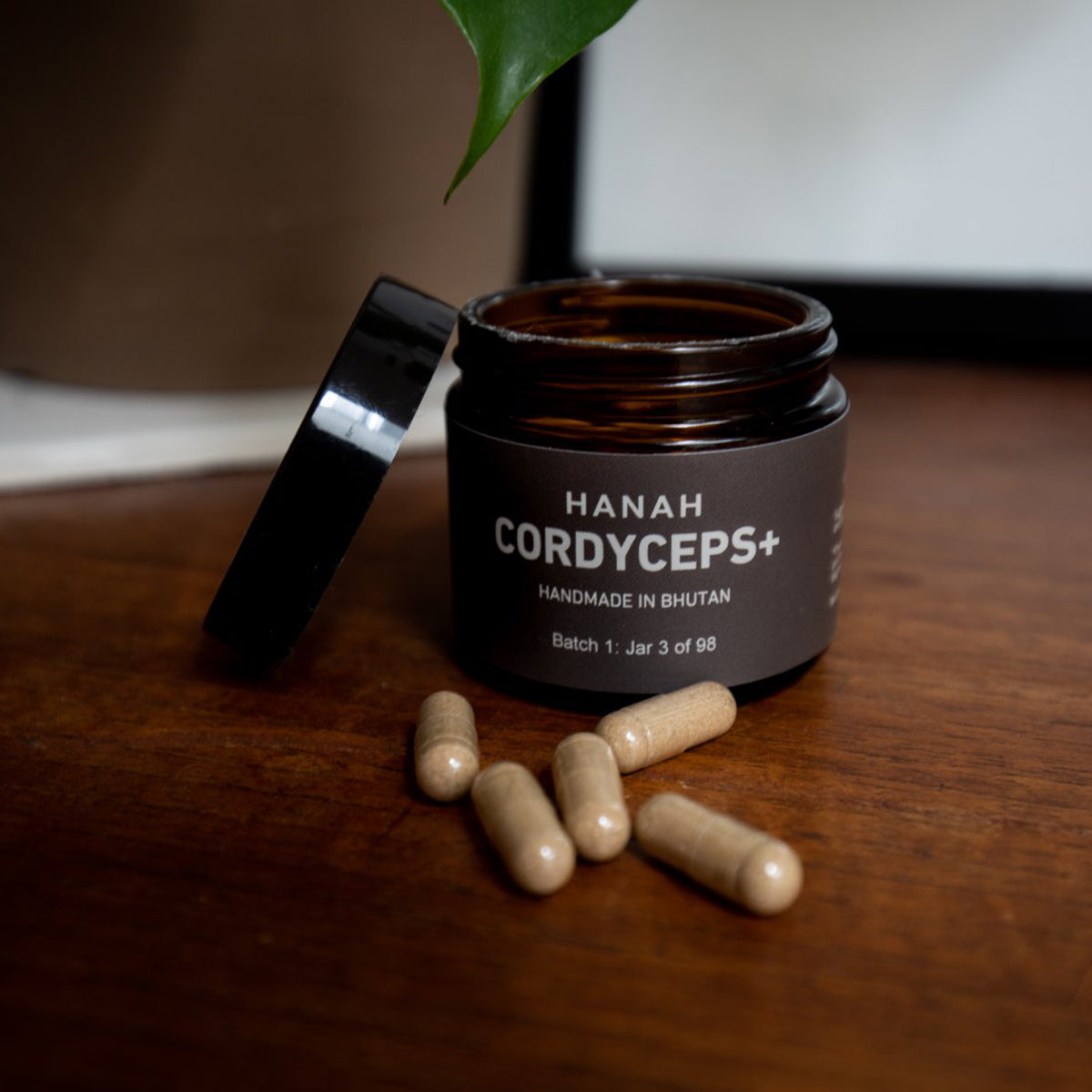 A jar of HANAH Cordyceps+ sits on a dark wood surface with the lid removed and leaning against the side of the jar and 5 capsules on the surface in front.